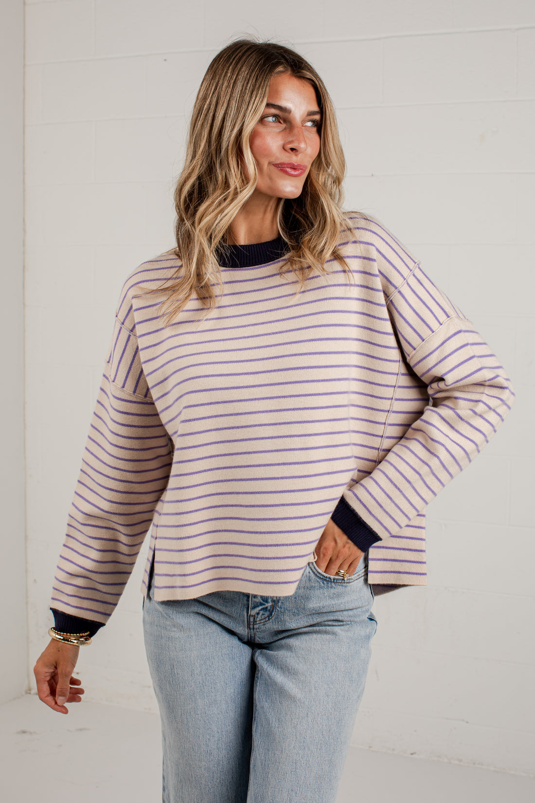 A woman wearing a long-sleeve, off-white ribbed knit sweater with thin purple and navy horizontal stripes, paired with light-wash blue jeans, standing against a white paneled wall.