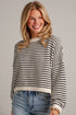 A smiling blonde woman wearing a cream and black horizontally striped knit sweater with long sleeves and blue jeans, standing against a light gray brick wall.