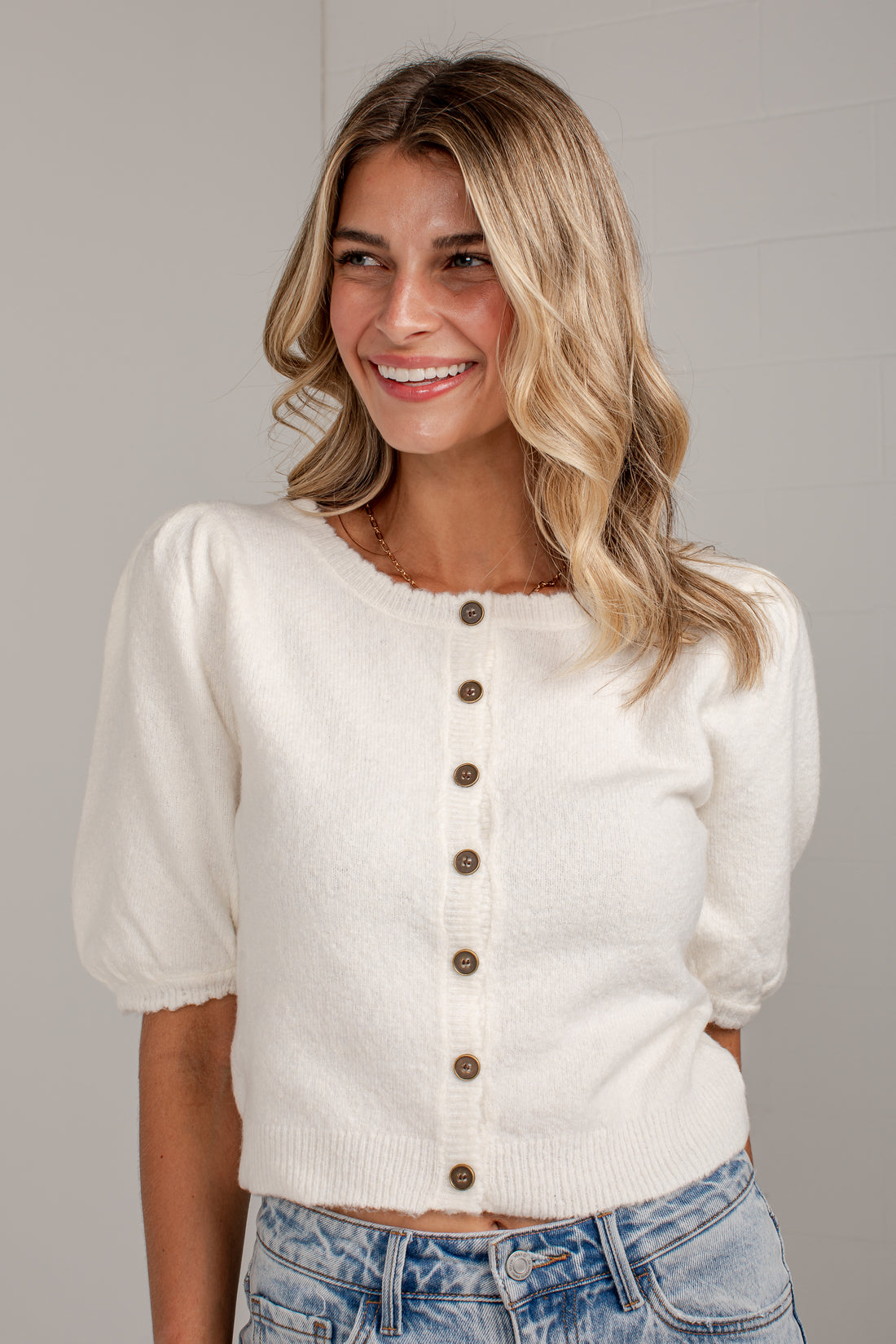 A smiling blonde woman wearing a cream-colored knit cardigan with brown buttons and puff sleeves, paired with light-wash blue jeans, against a neutral gray background.