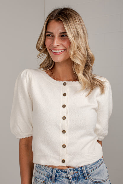 A smiling blonde woman wearing a cream-colored knit cardigan with brown buttons and puff sleeves, paired with light-wash blue jeans, against a neutral gray background.