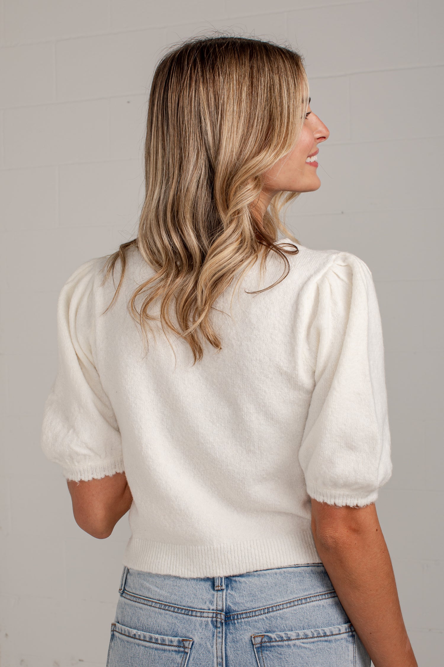 A woman with blonde-highlighted hair is shown from the back wearing a cream puff-sleeve knit top and light blue jeans against a light gray wall.