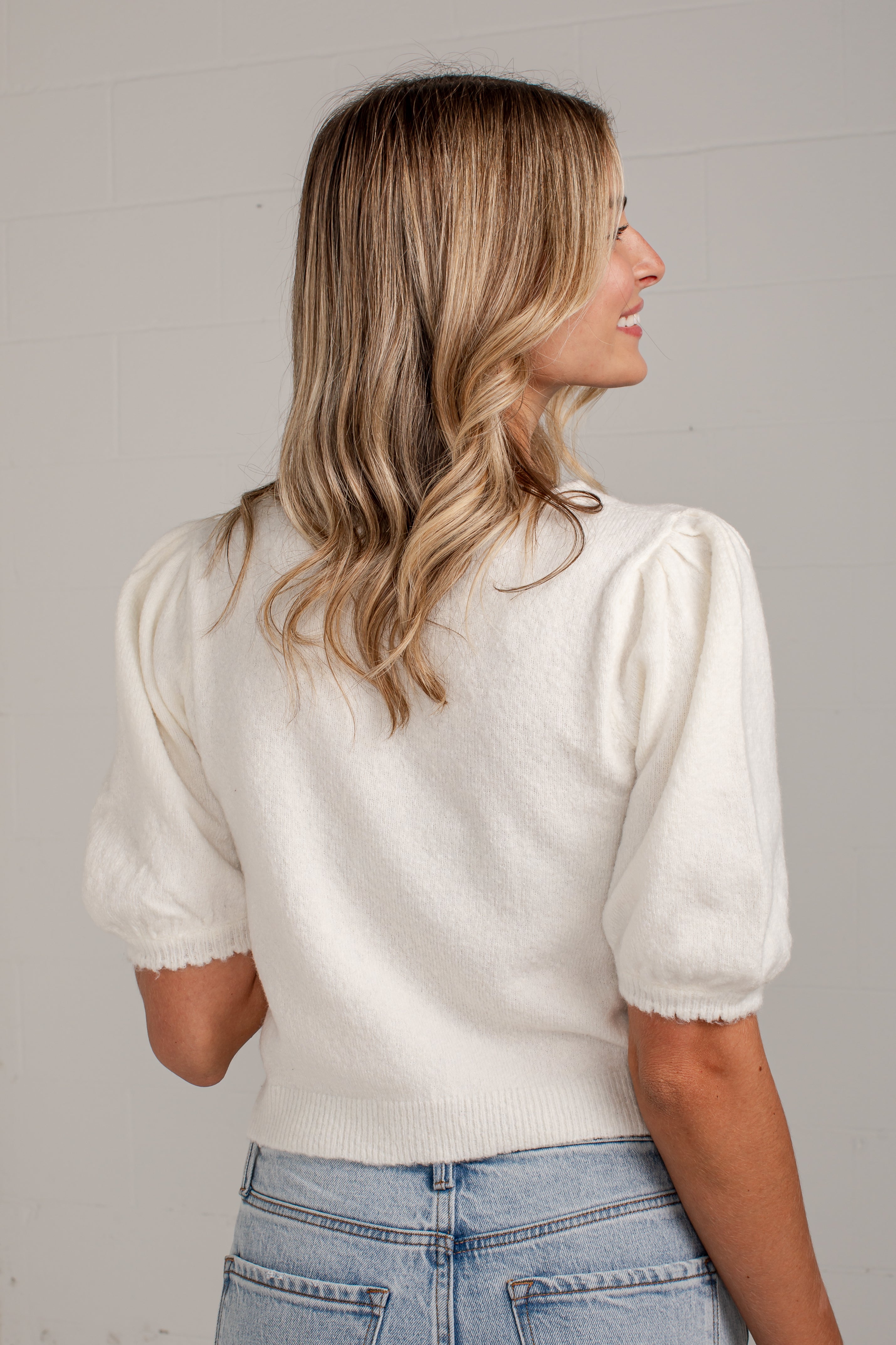 A woman with blonde-highlighted hair is shown from the back wearing a cream puff-sleeve knit top and light blue jeans against a light gray wall.