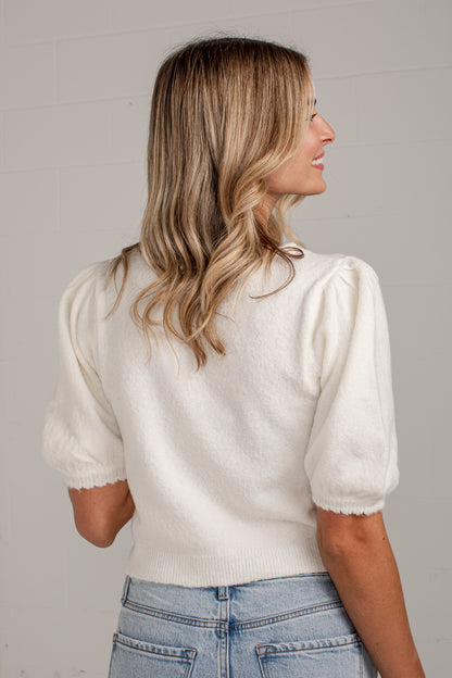 A woman with blonde-highlighted hair is shown from the back wearing a cream puff-sleeve knit top and light blue jeans against a light gray wall.