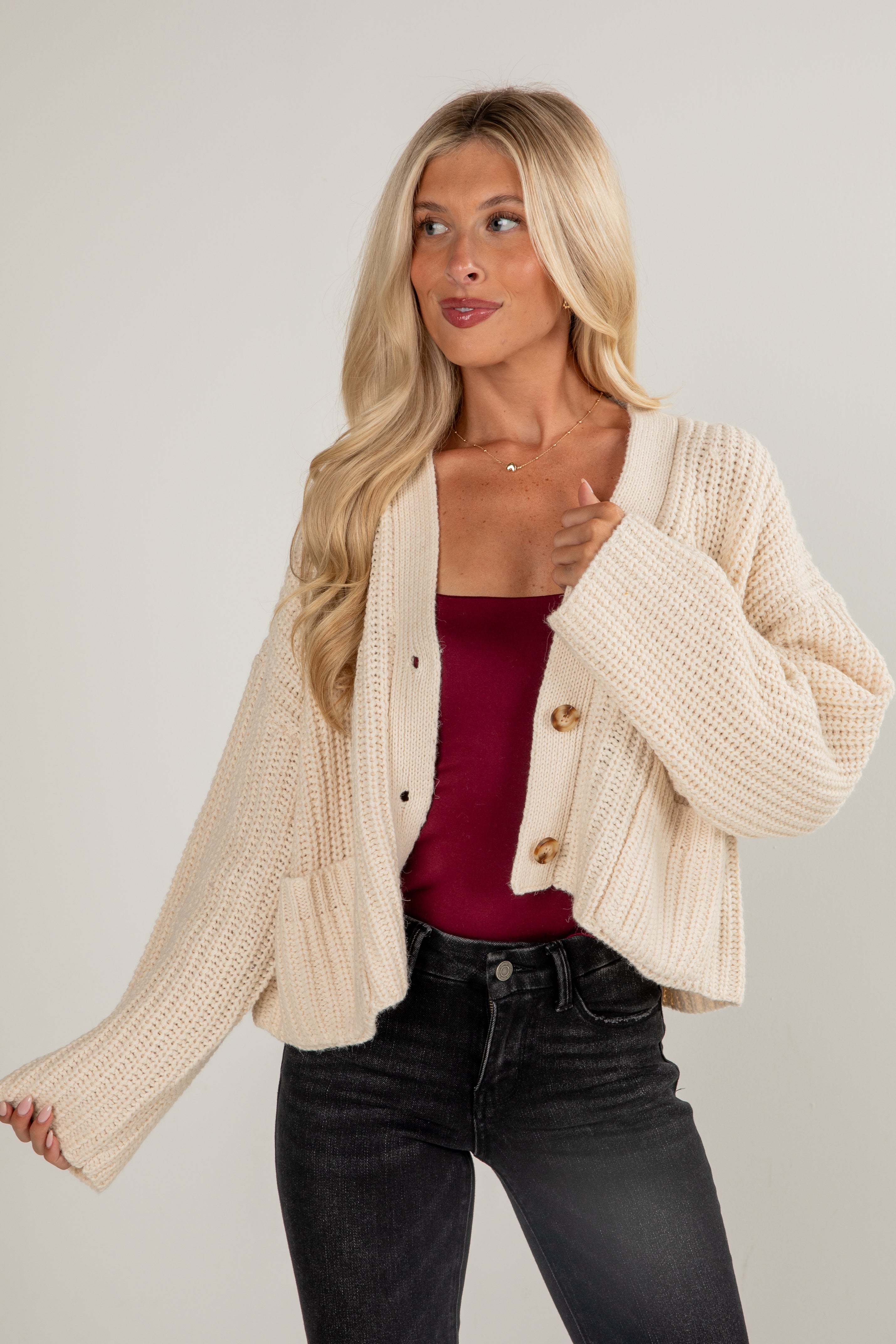 Woman wearing a beige knitted cardigan over a red top and black jeans on a plain background