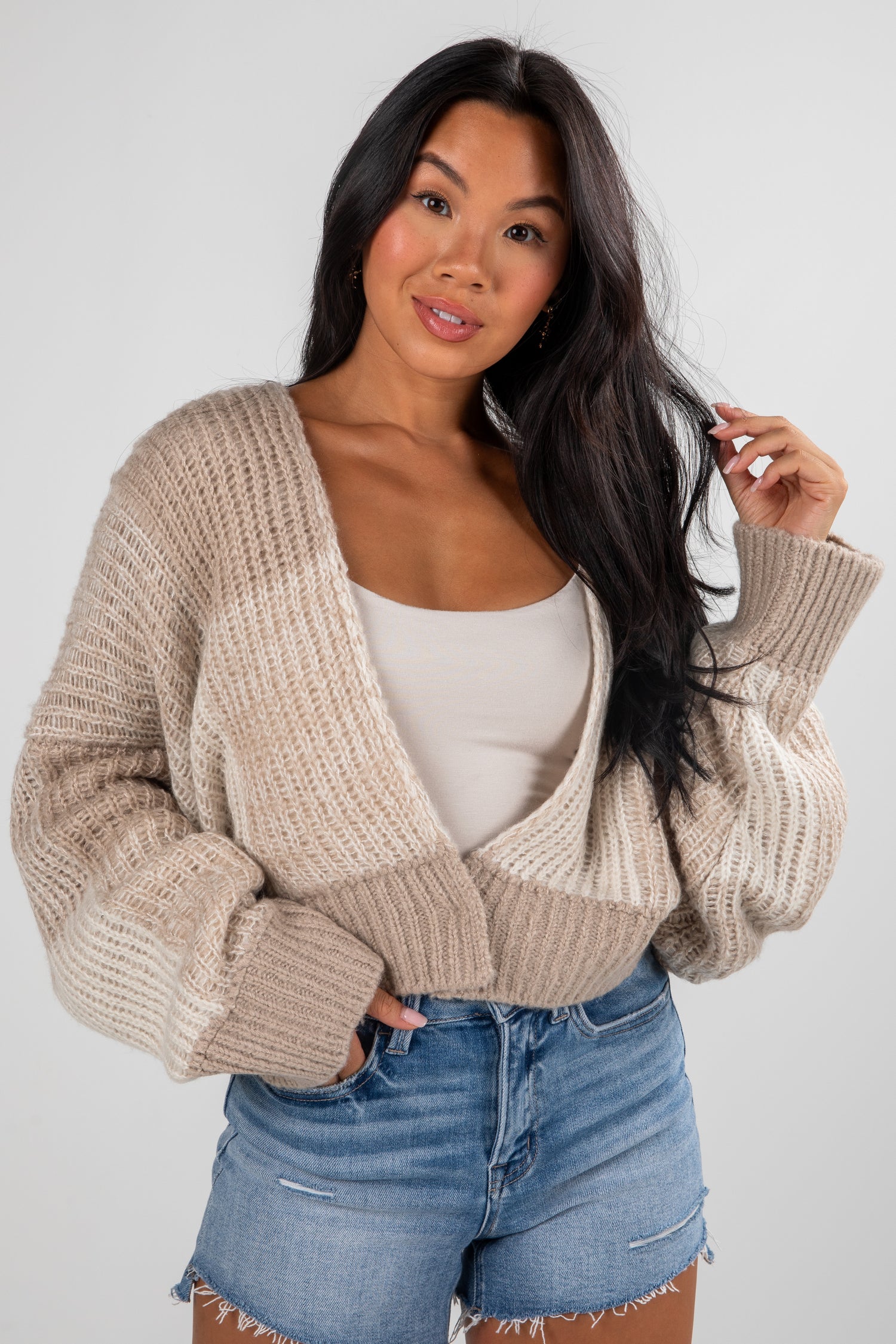 Woman wearing a beige knitted cardigan over a white top and blue shorts on a plain background