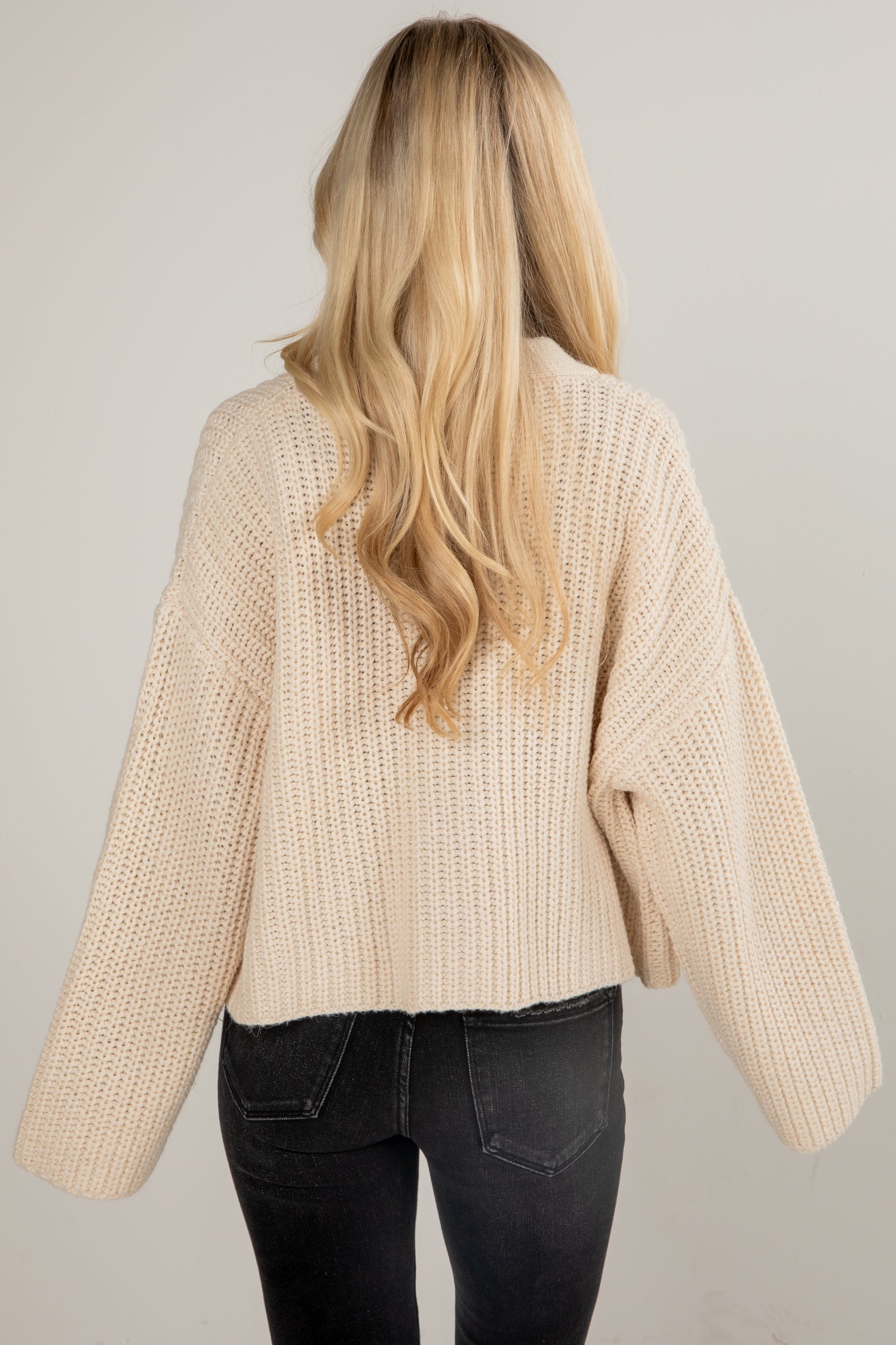 Person wearing a beige knitted sweater and black pants on a plain background