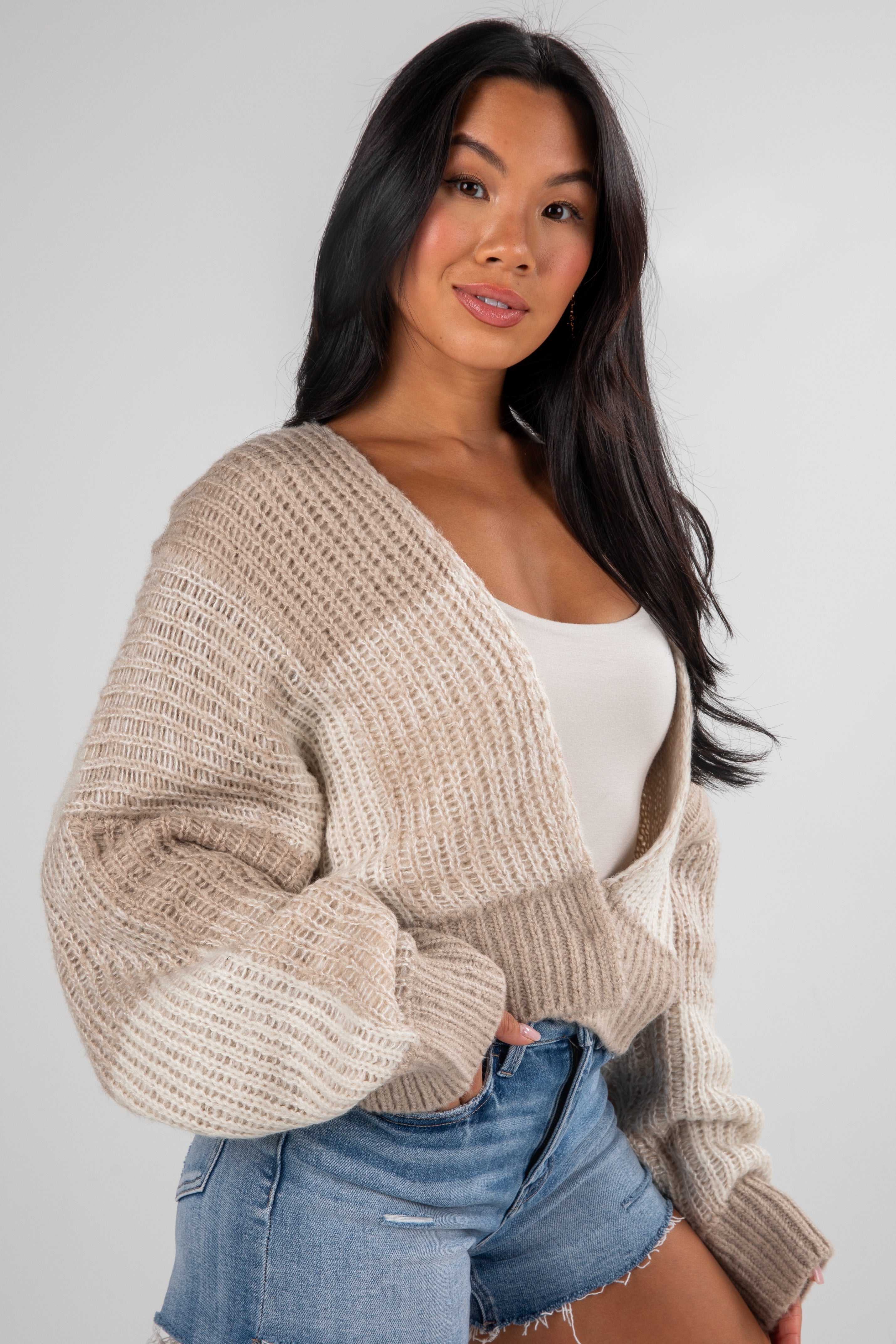 Woman wearing a beige knitted cardigan over a white top and denim shorts on a plain background