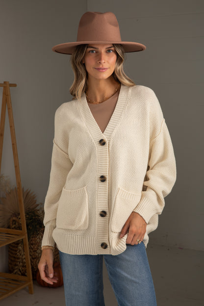 Woman wearing a beige cardigan, brown hat, and blue jeans against a plain background