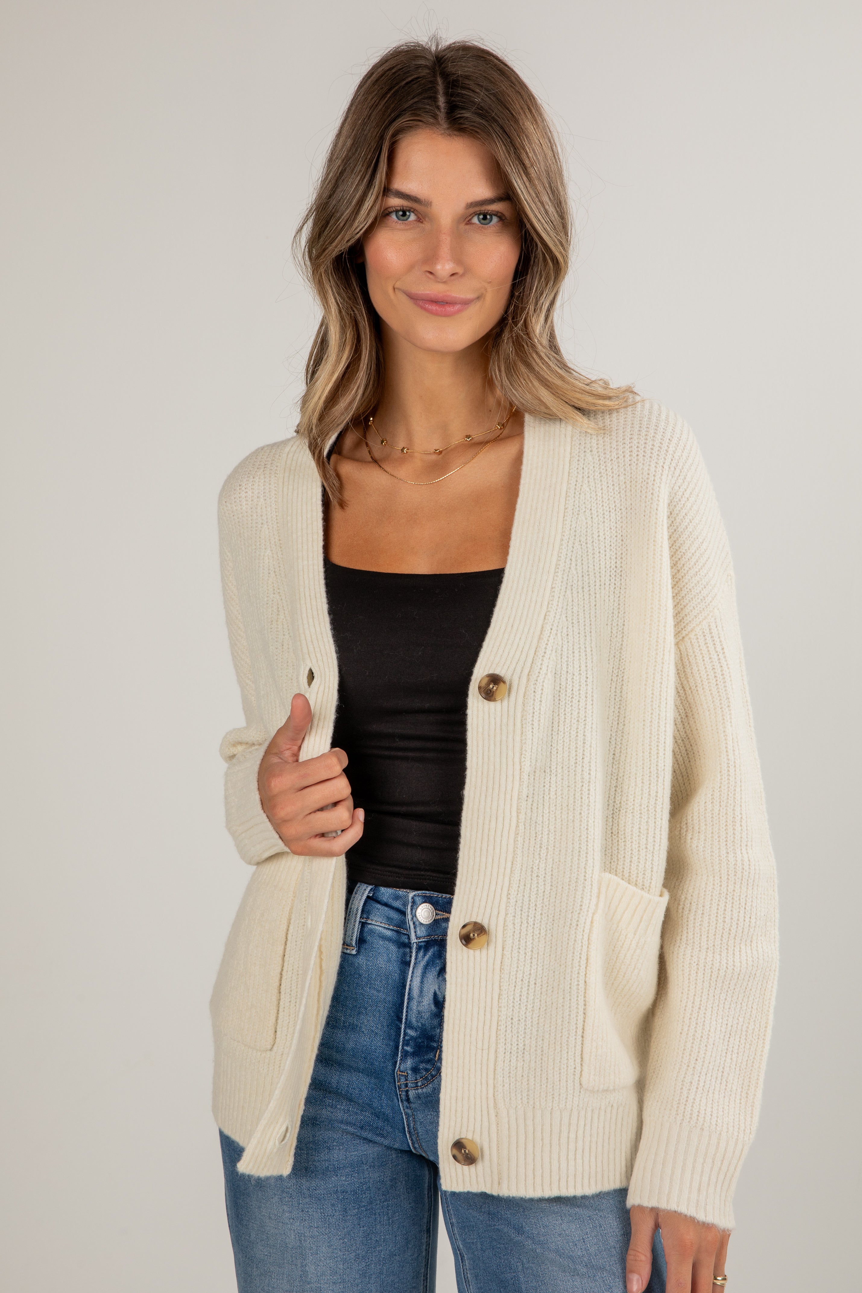 Woman wearing a cream cardigan over a black top and blue jeans on a plain background