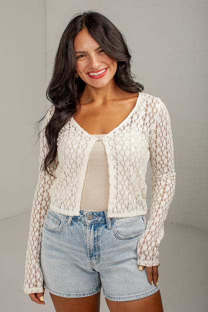 A woman modeling a cream-colored, long-sleeve crochet knit cardigan over a beige top and light-wash denim shorts, standing against a neutral light gray studio background.