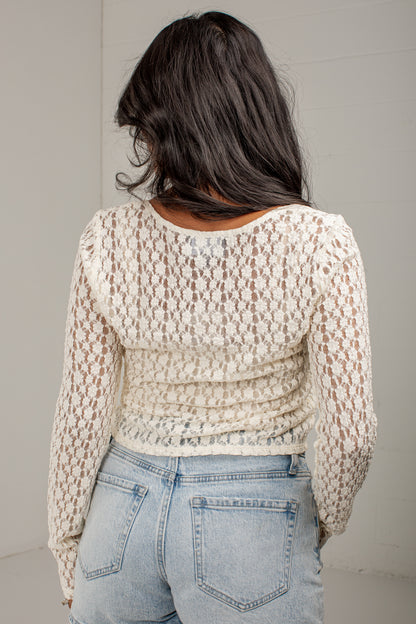 A woman viewed from behind wearing a cream-colored, long-sleeve open-knit sweater and light-wash blue jeans against a plain light gray wall.