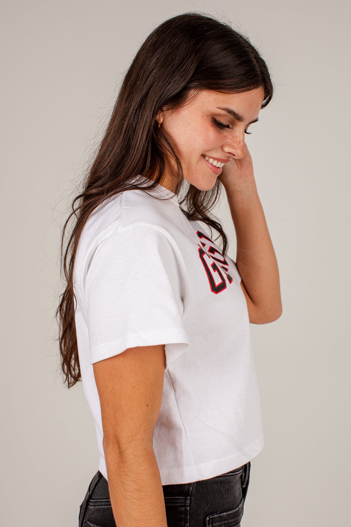 A woman with long dark brown hair is shown in profile, wearing a white short-sleeve crop top with a red and black graphic on the chest, paired with black jeans, against a plain light gray background.