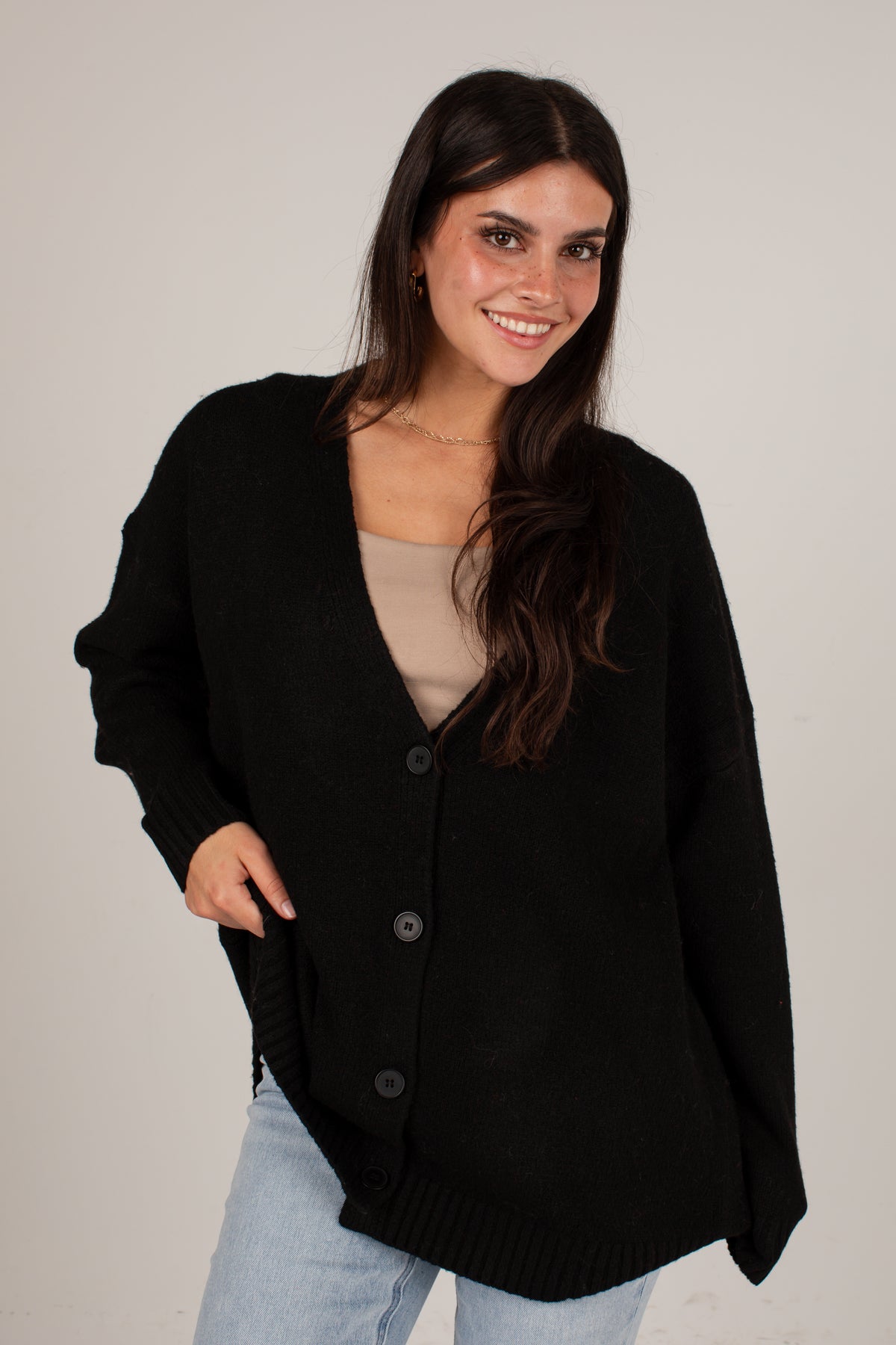 A woman modeling an oversized black knit cardigan with a V-neck and front buttons, worn over a beige top and light blue jeans, against a plain light gray studio background.