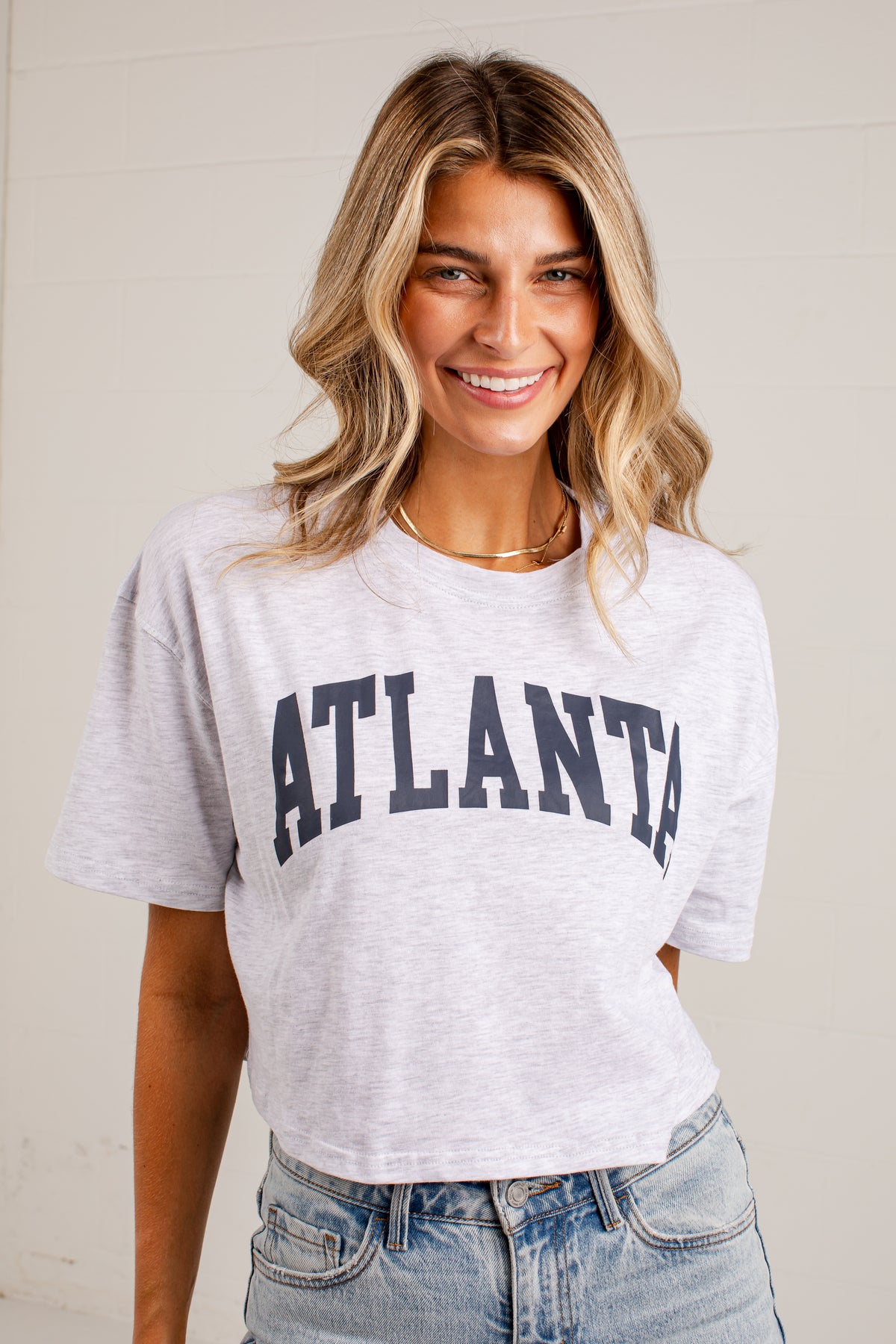 A smiling woman with blonde-highlighted hair wearing a light heather gray cropped T-shirt with &