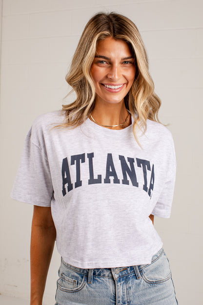 A smiling woman with blonde-highlighted hair wearing a light heather gray cropped T-shirt with &