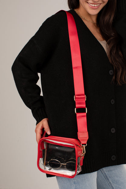 A woman wearing a black cardigan and light blue jeans carries a small transparent waterproof pouch bag with a bright red strap and red trim. The pouch contains round sunglasses.
