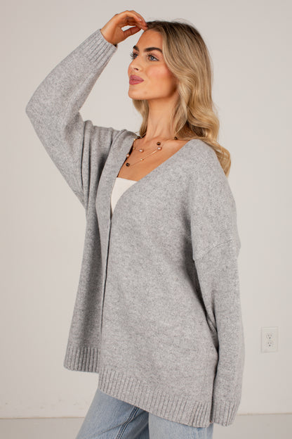 A woman wearing an oversized light gray knit cardigan over a white top and light blue jeans, standing against a plain off-white wall.