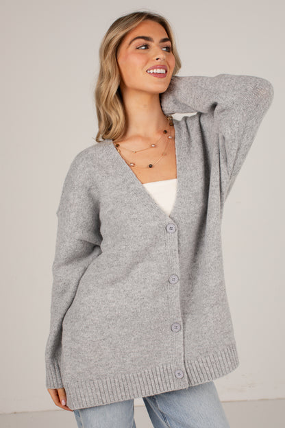 A woman wearing an oversized light gray knit cardigan with a V-neck and front buttons, layered over a white top and light blue jeans, photographed against a plain light gray studio background.