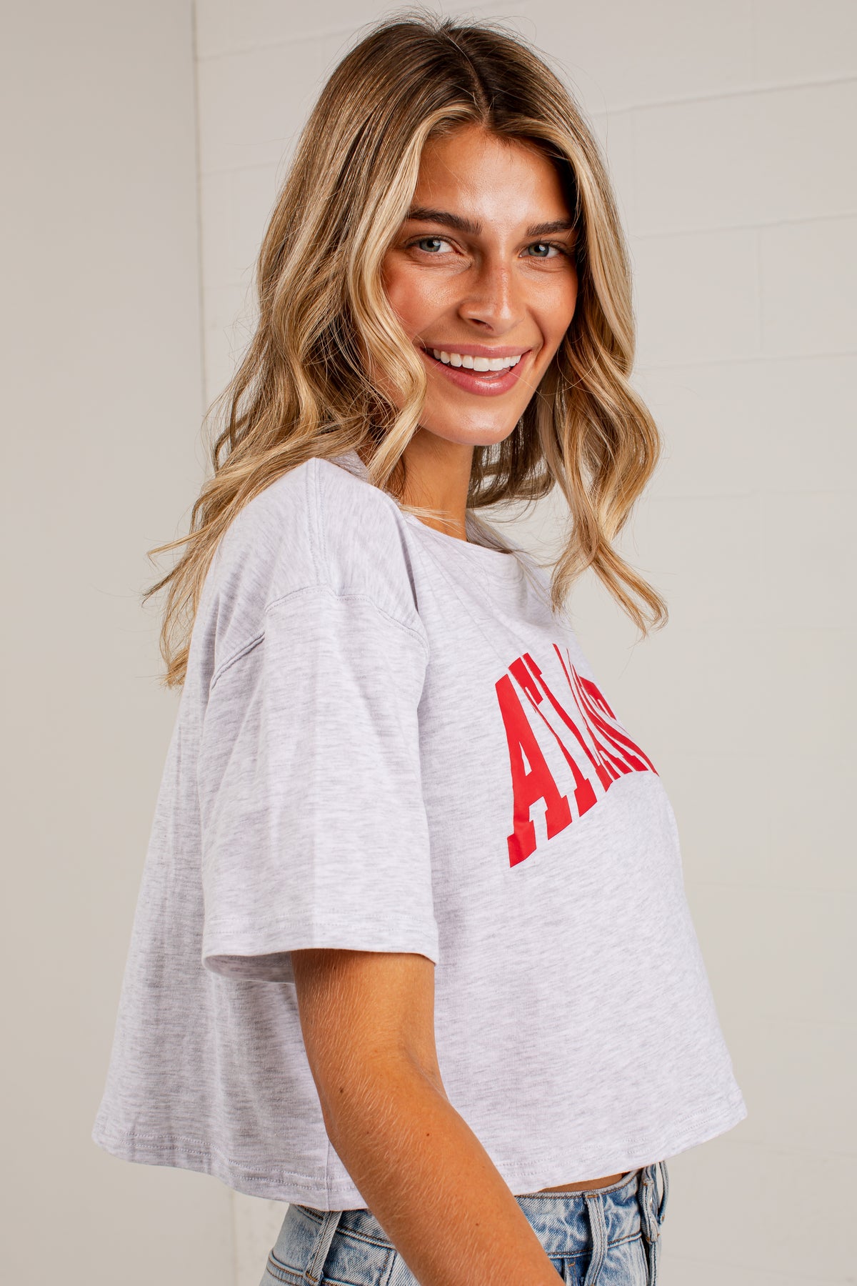 A woman wearing a light heather gray cropped t-shirt with a bold red &