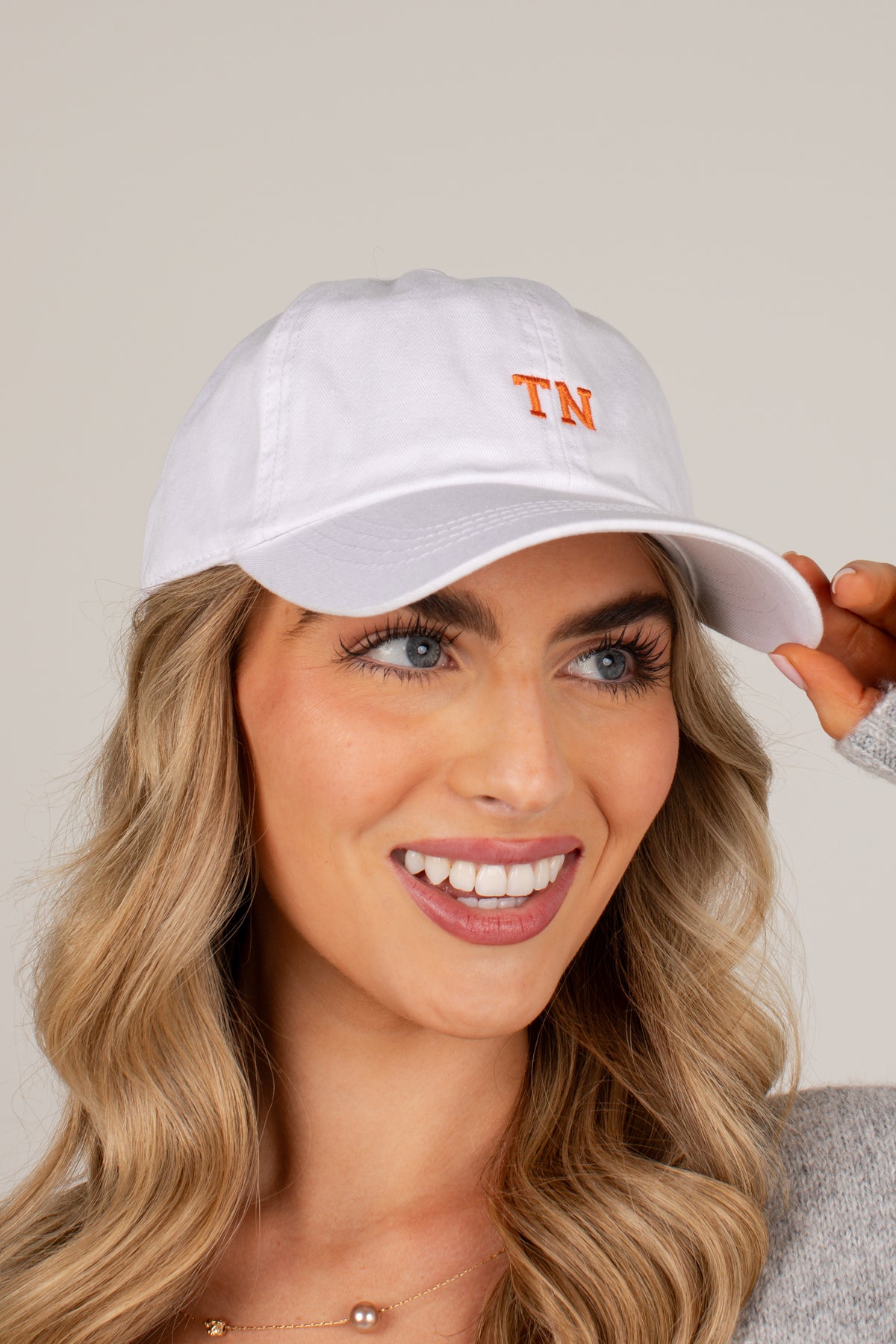 A woman with blonde wavy hair wearing a white baseball cap with orange embroidered &