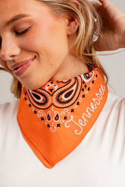 A close-up of a woman wearing an orange bandana-style neck gaiter with a black, white, and brown paisley pattern and the word &