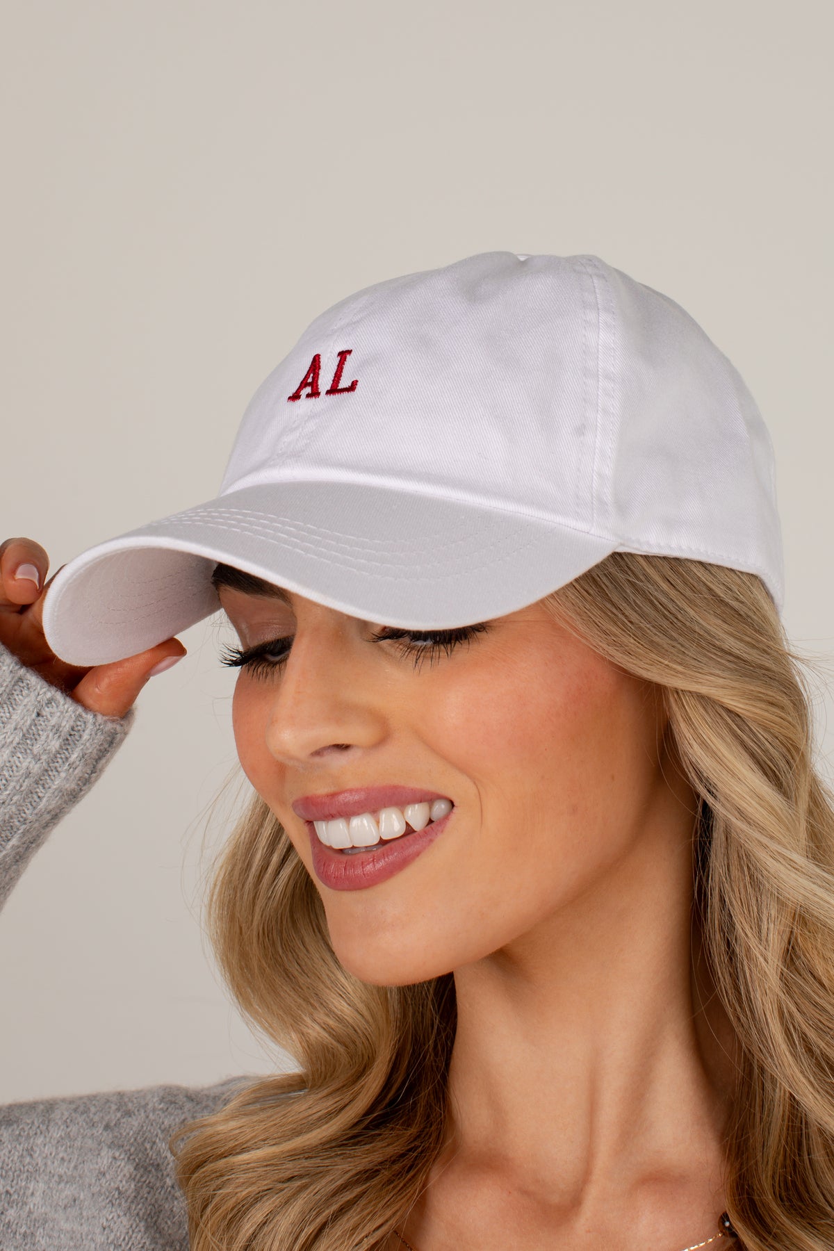 A close-up of a smiling blonde woman wearing a white baseball cap with red embroidered letters &