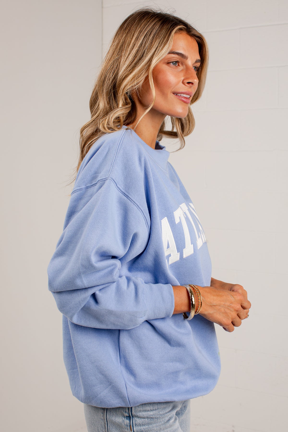 A woman wearing a light blue crewneck sweatshirt with white varsity-style lettering on the front, paired with light-wash jeans, photographed against a white paneled wall.