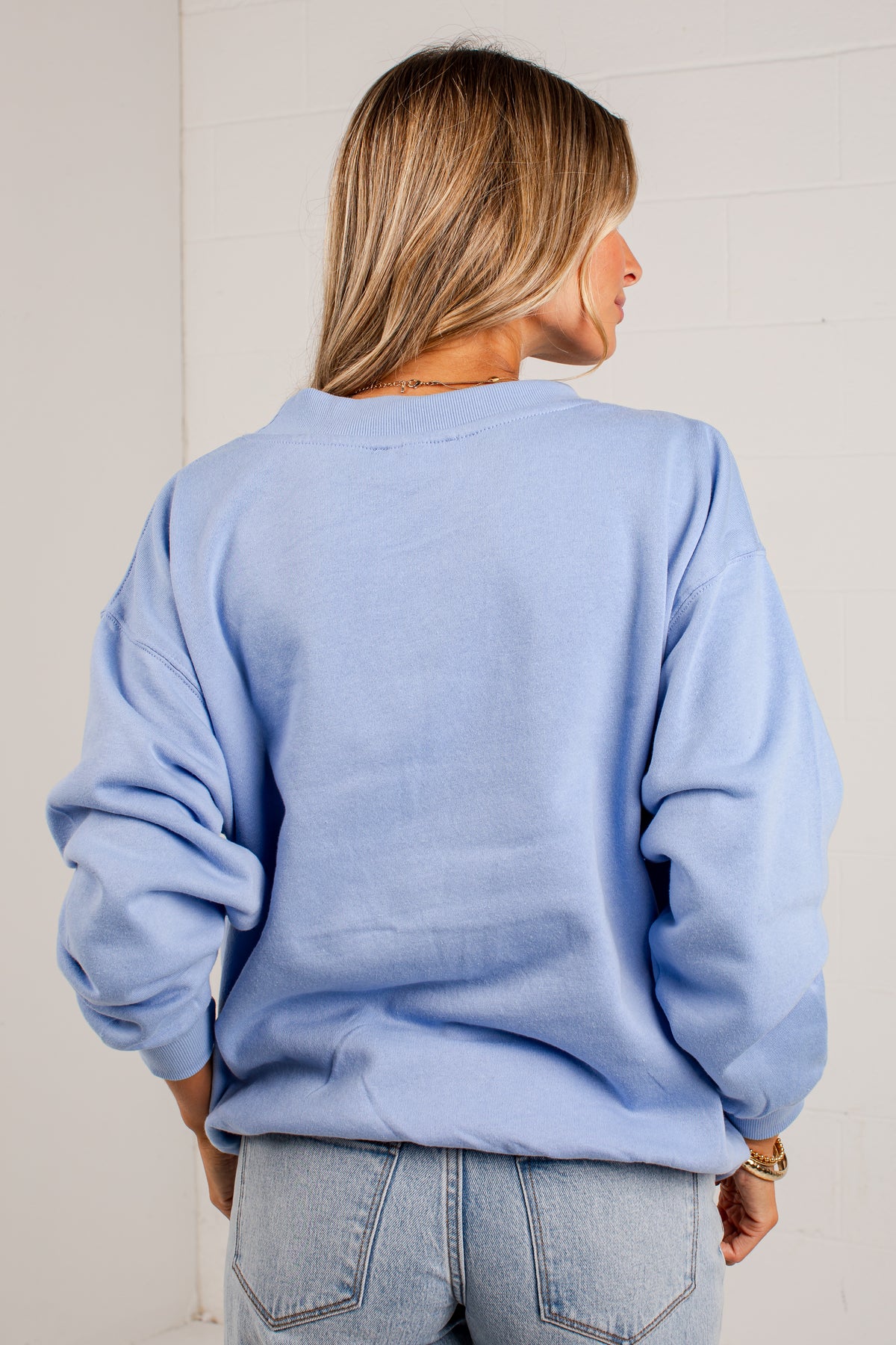 A woman viewed from behind wearing an oversized light blue crewneck sweatshirt and light-wash jeans against a white paneled wall.