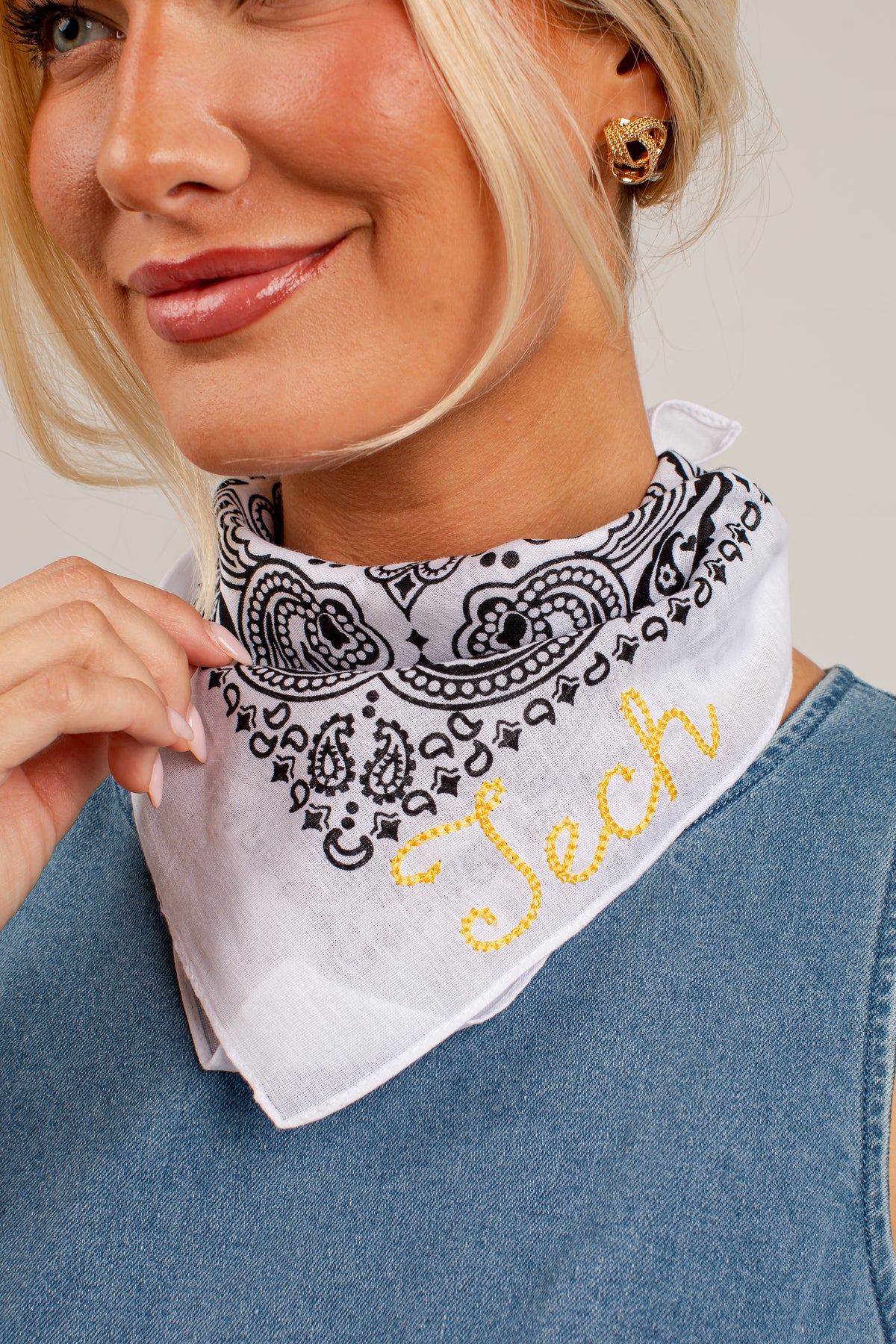 A close-up of a woman wearing a white bandana-style neck gaiter with a black paisley pattern and yellow embroidered script reading &