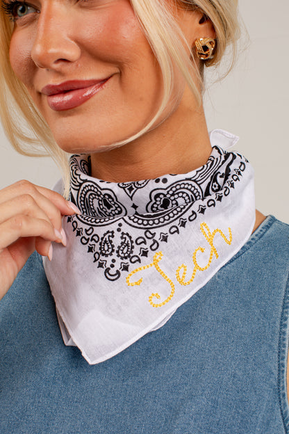 A close-up of a woman wearing a white bandana-style neck gaiter with a black paisley pattern and yellow embroidered script reading &