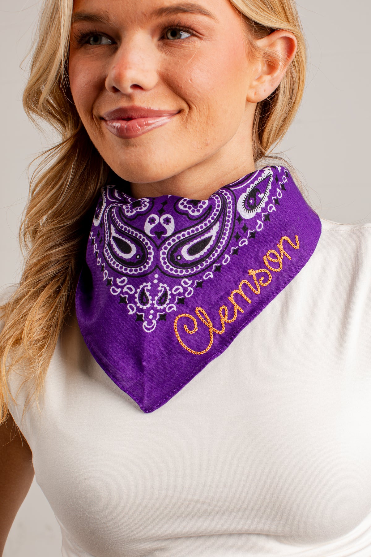 A close-up of a woman wearing a purple bandana-style neck gaiter with white paisley embroidery and the name &