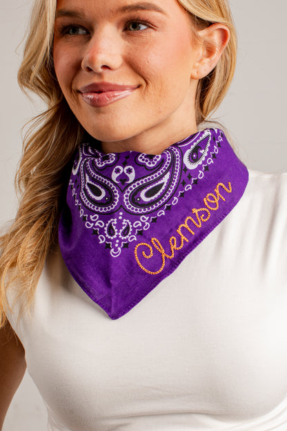 A close-up of a woman wearing a purple bandana-style neck gaiter with white paisley embroidery and the name &
