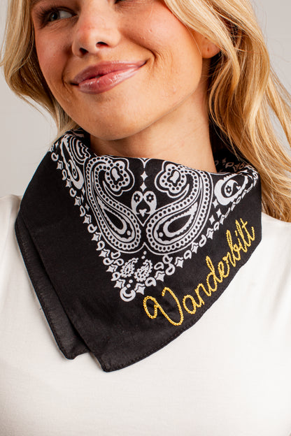 A close-up of a woman wearing a black bandana-style neck gaiter with a white paisley pattern and gold embroidered text reading &