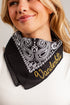 A close-up of a woman wearing a black bandana-style neck gaiter with a white paisley pattern and gold embroidered text reading &
