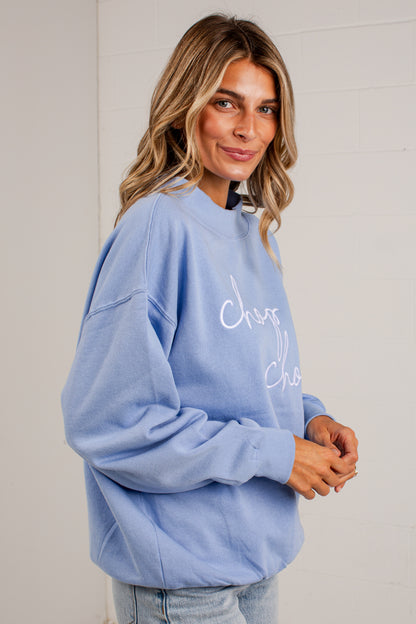 A woman wearing a light blue crewneck sweatshirt with white cursive text on the front, paired with light wash jeans, standing against a white brick wall.