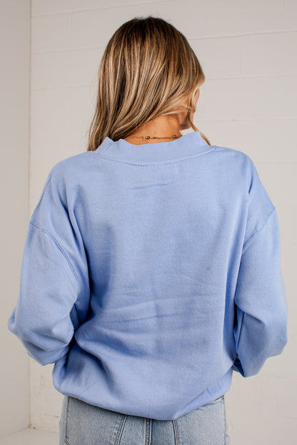 A woman viewed from behind wearing a light blue crewneck sweatshirt and light wash jeans, standing against a white paneled wall.