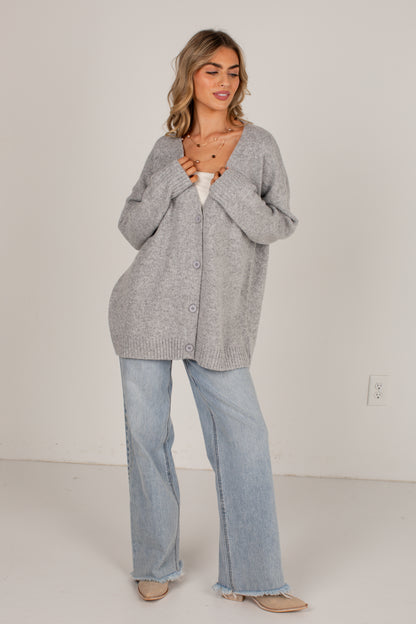 A woman modeling an oversized light gray knit cardigan with front buttons, worn over a white top and paired with light blue wide-leg jeans and beige flats, standing against a plain off-white wall.