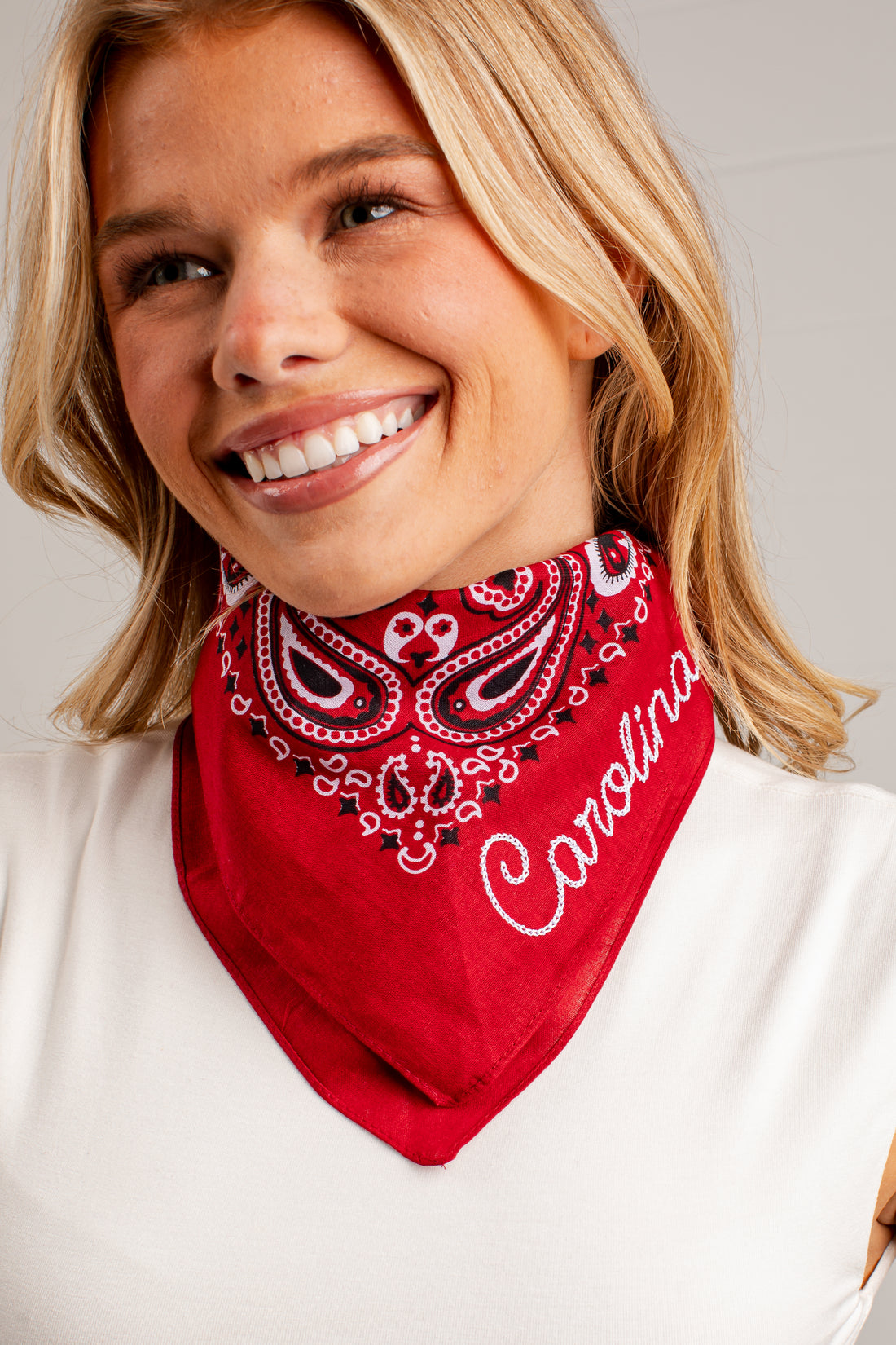 A smiling blonde woman wearing a red bandana-style neck gaiter with a white paisley pattern and the word &