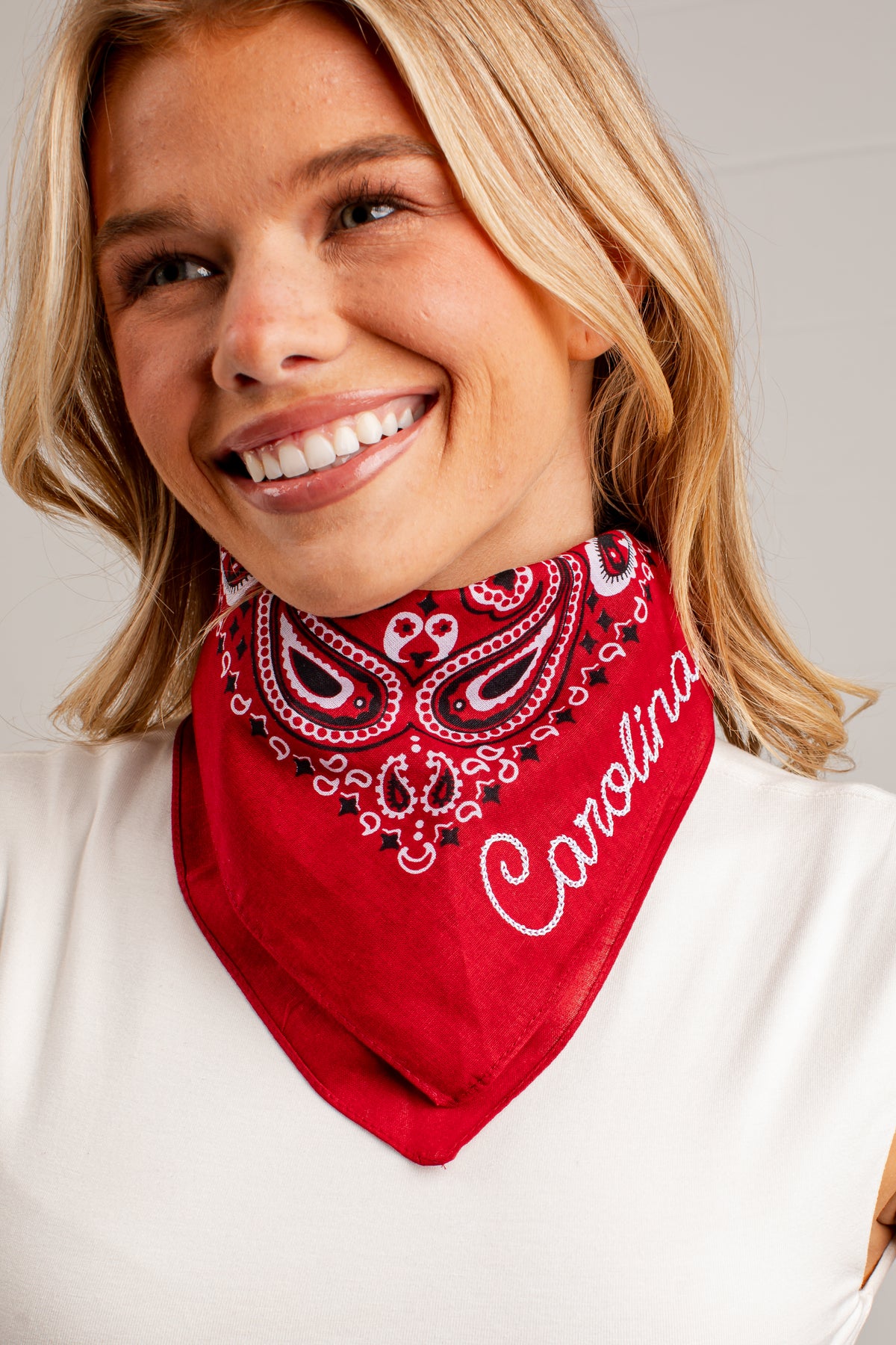 A smiling blonde woman wearing a red bandana-style neck gaiter with a white paisley pattern and the word &