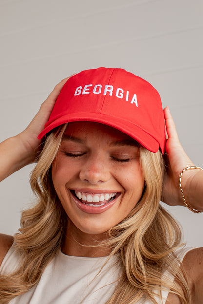 A smiling blonde woman wearing a bright red baseball cap with &