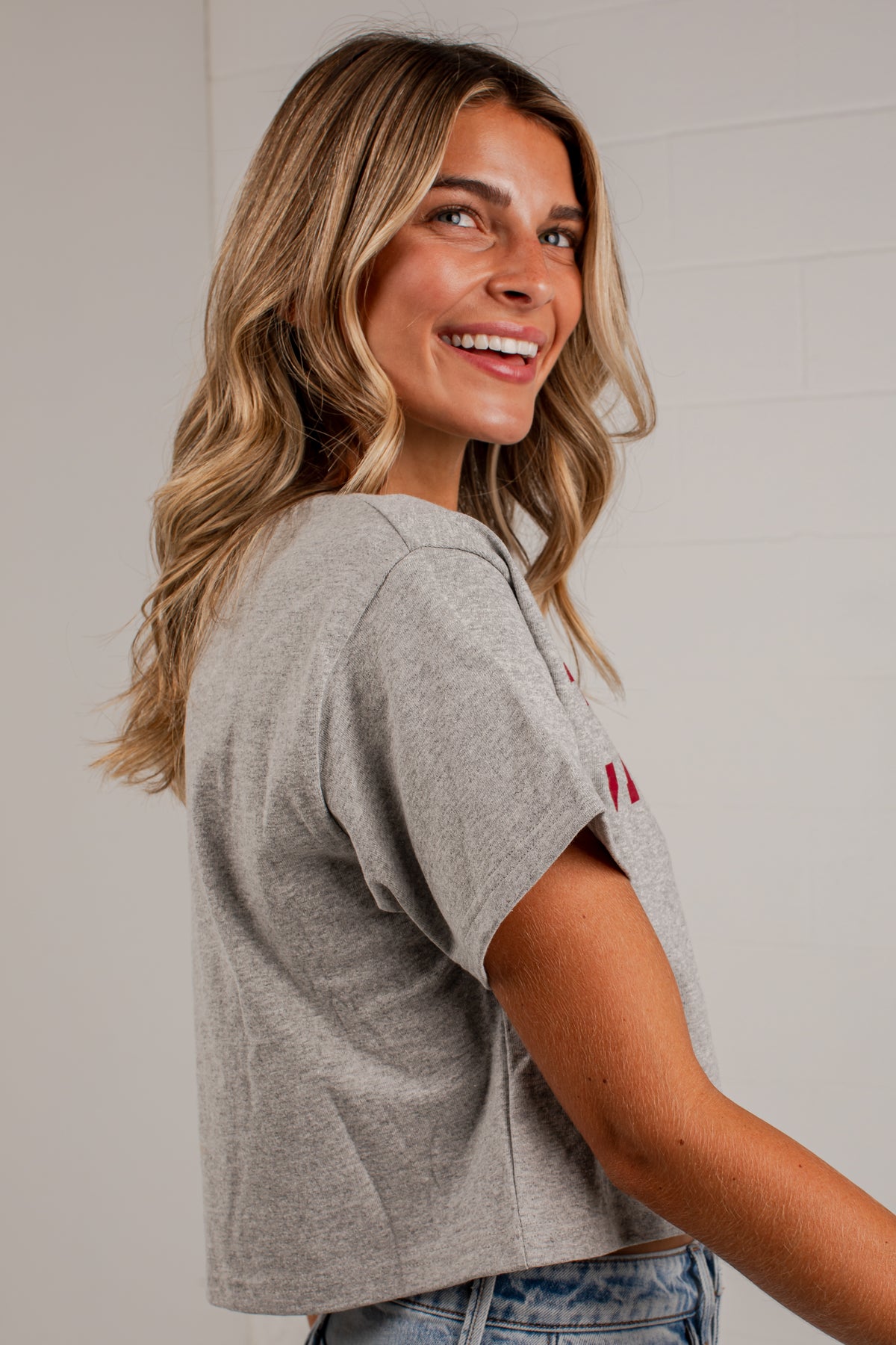 A woman with blonde wavy hair smiling over her shoulder, wearing a light gray heather cropped short-sleeve t-shirt with a small red embroidered logo on the sleeve, paired with blue jeans against a white brick wall background.