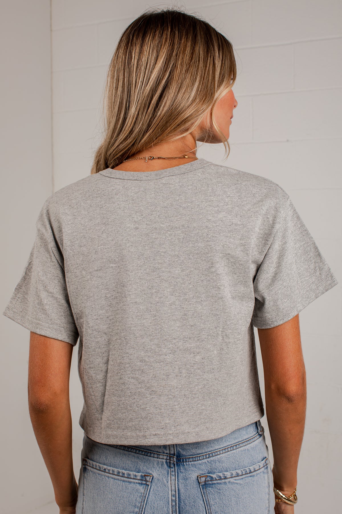 A woman viewed from the back wearing a light heather gray short-sleeve crop top and light blue jeans, standing against a white paneled wall.