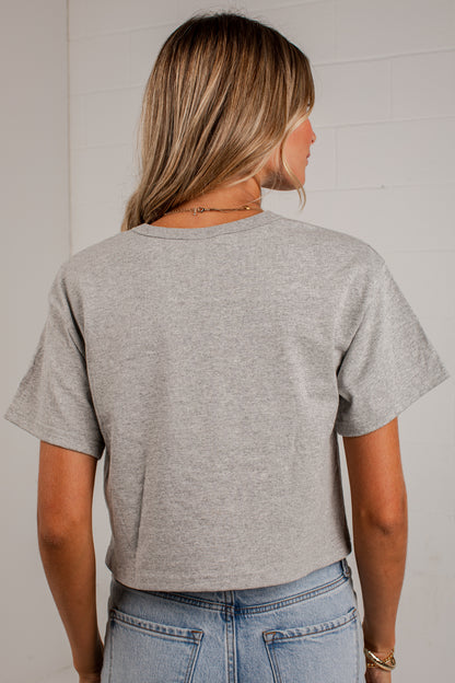 A woman viewed from the back wearing a light heather gray short-sleeve crop top and light blue jeans, standing against a white paneled wall.