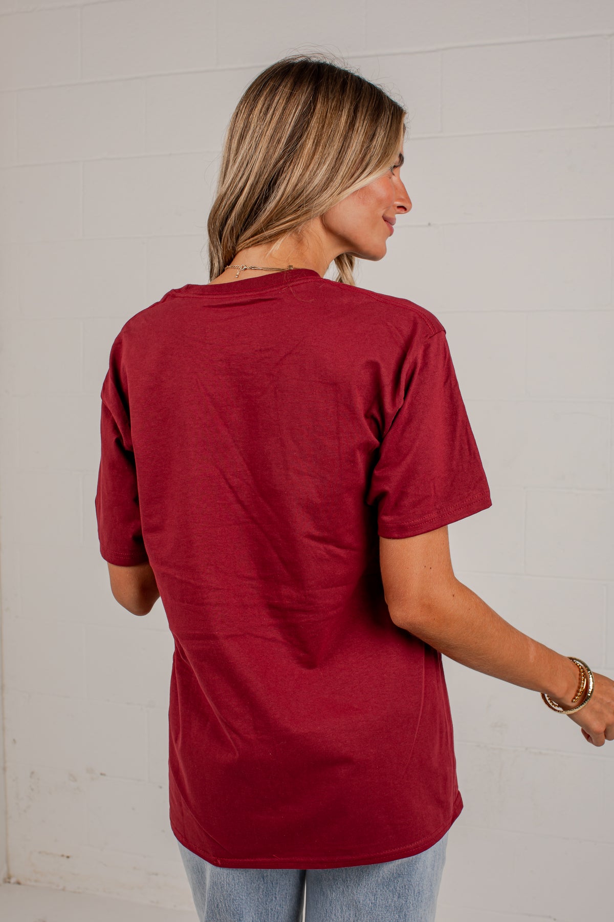 A woman with blonde hair is shown from behind wearing a short-sleeve maroon t-shirt and light blue jeans, standing against a white brick wall.