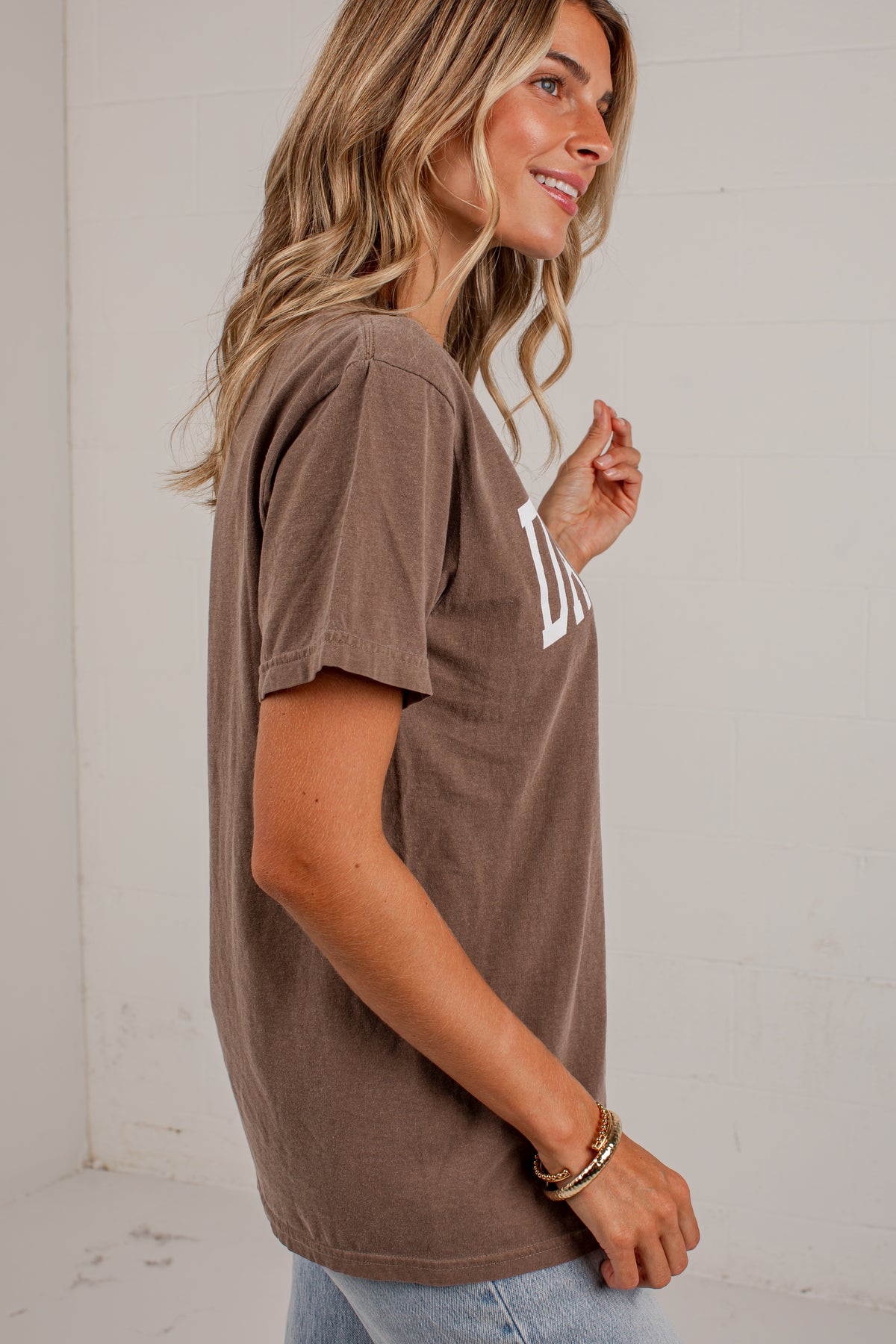 A woman wearing a brown short-sleeve t-shirt with a white graphic print on the chest, paired with light blue jeans, standing against a white brick wall.