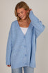 A woman wearing an oversized light blue knit cardigan with a white V-neck undershirt and light blue jeans, photographed against a plain light beige studio background.