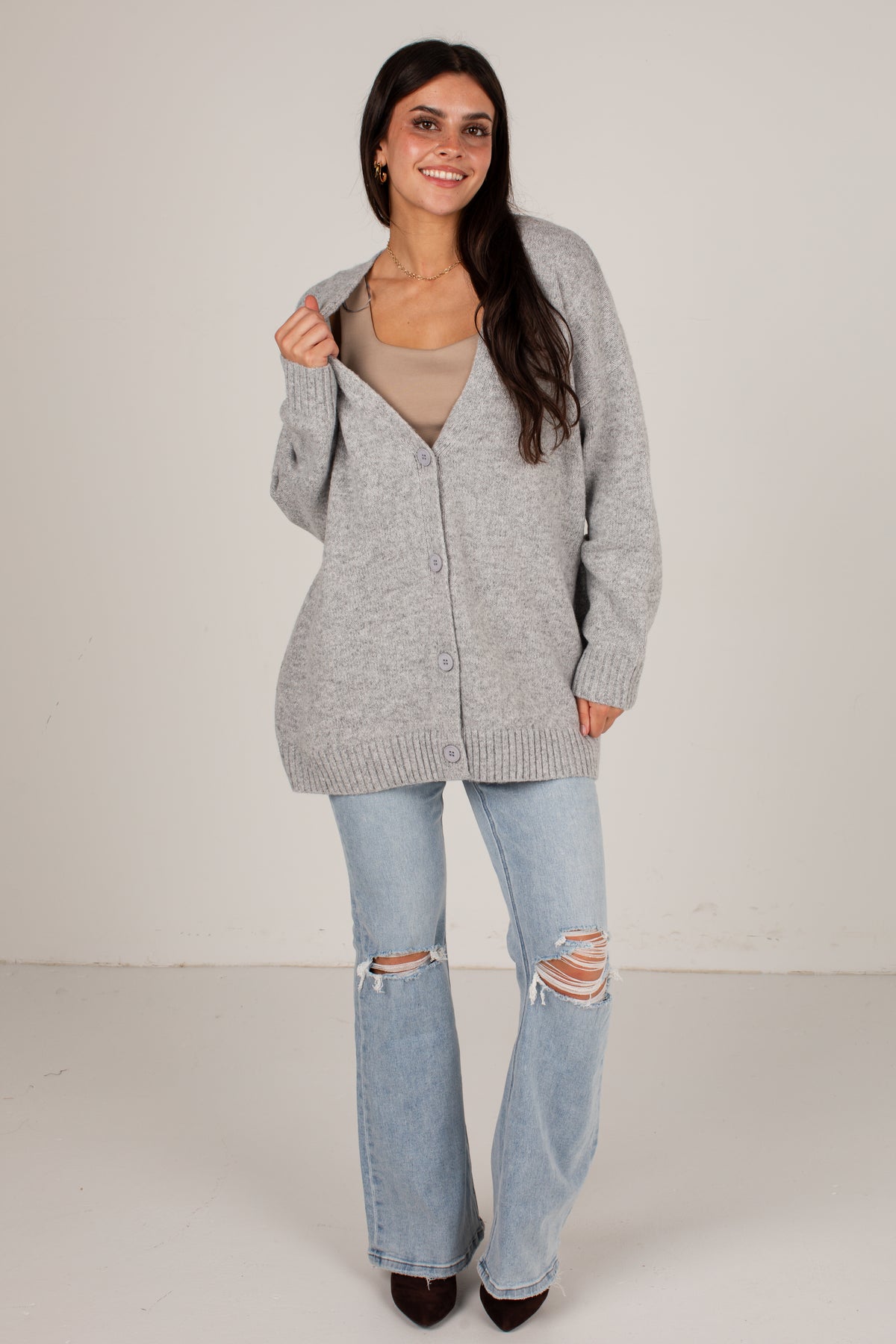 A woman modeling a light gray button-up knit cardigan over a beige top and light-wash distressed flare jeans, standing against a plain off-white studio background.