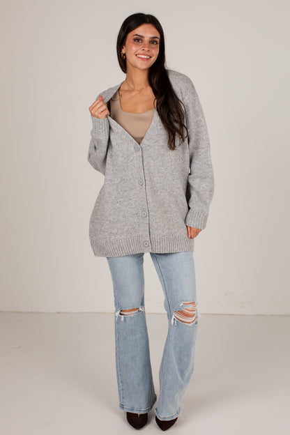 A woman modeling a light gray button-up knit cardigan over a beige top and light-wash distressed flare jeans, standing against a plain off-white studio background.