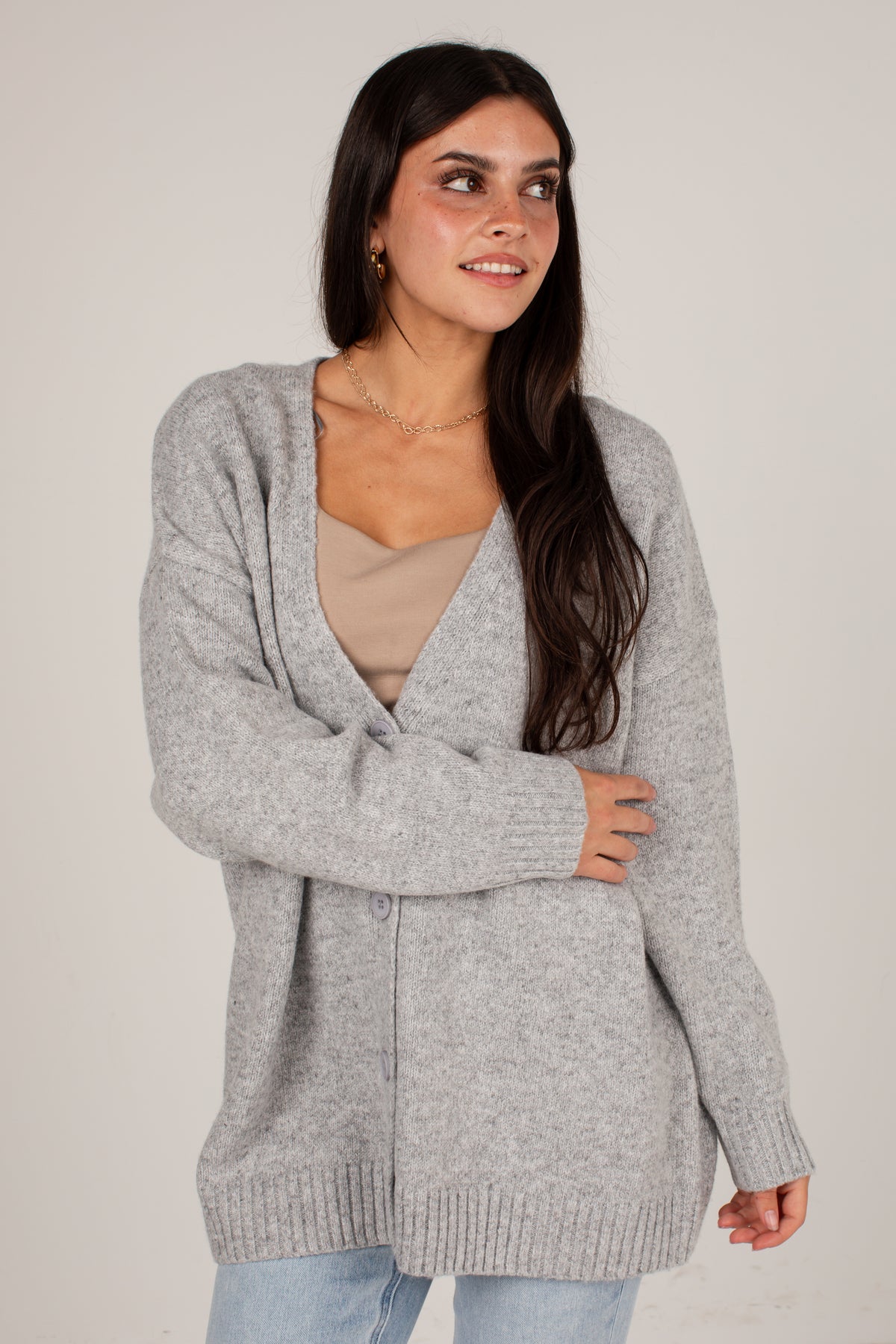 A woman wearing a light gray knit cardigan over a beige top and light blue jeans, standing against a plain light gray studio background.