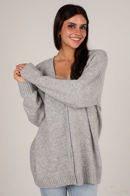 A woman modeling an oversized light gray knit cardigan sweater with a V-neck and ribbed cuffs and hem, paired with light blue jeans against a plain light gray studio background.